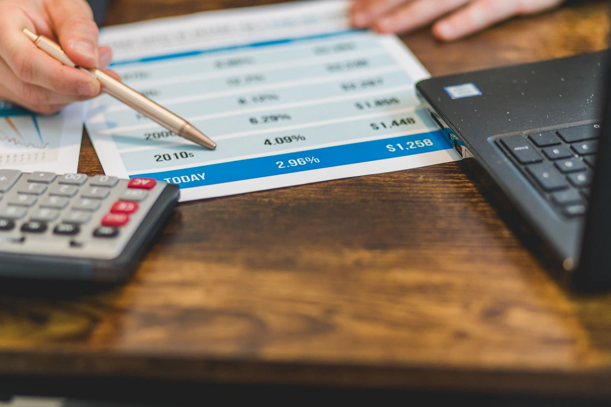 Hand holding a pen over printed financial figures next to a calculator and laptop, working through runway calculations