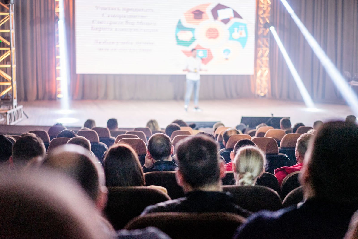 Audience seated in a dark auditorium watching a speaker on a lit conference stage