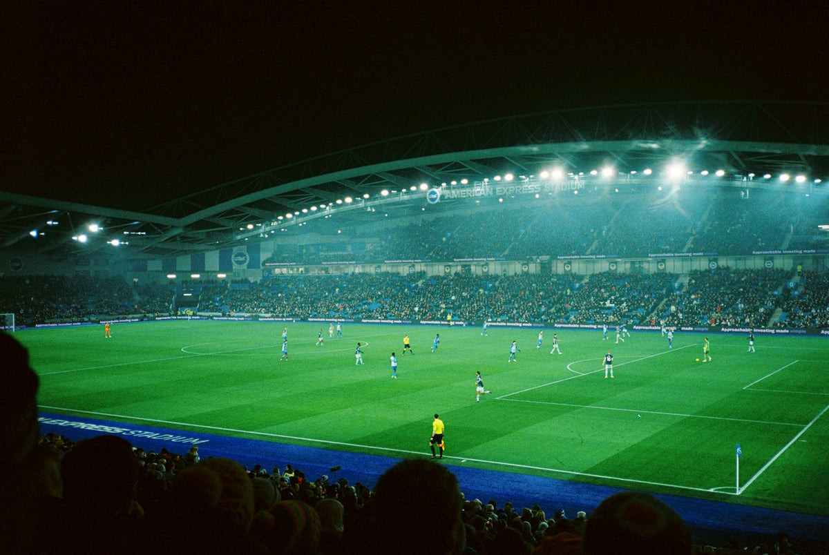 Floodlit football stadium at night during a match, seen from the stands